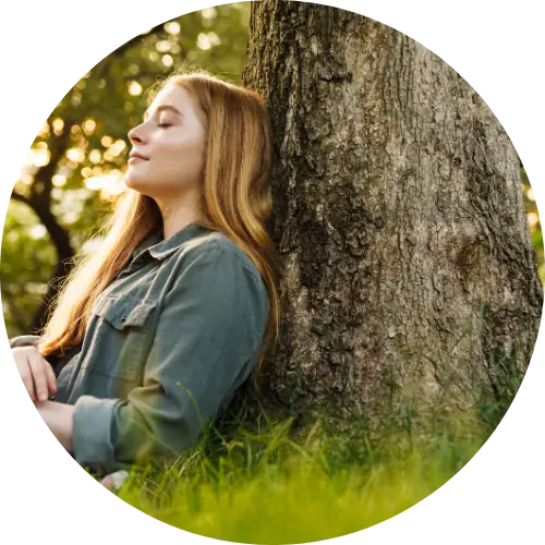Young woman with long auburn hair leaning against a large tree trunk in grass, eyes closed, face tilted up calmly.