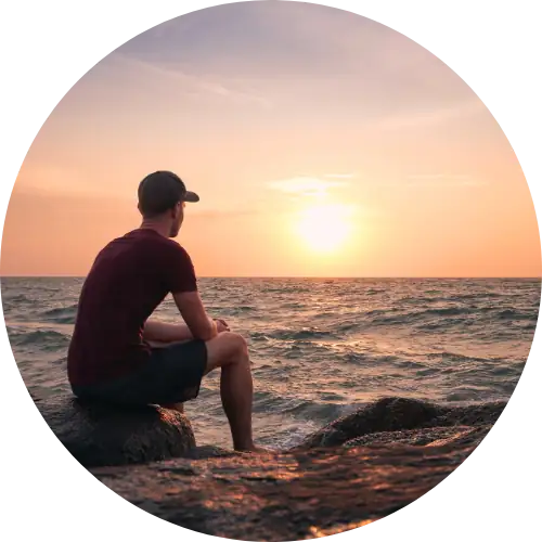 A young man in a red t-shirt and cap sits on a rock at the sea edge, watching a low sun set over the water.